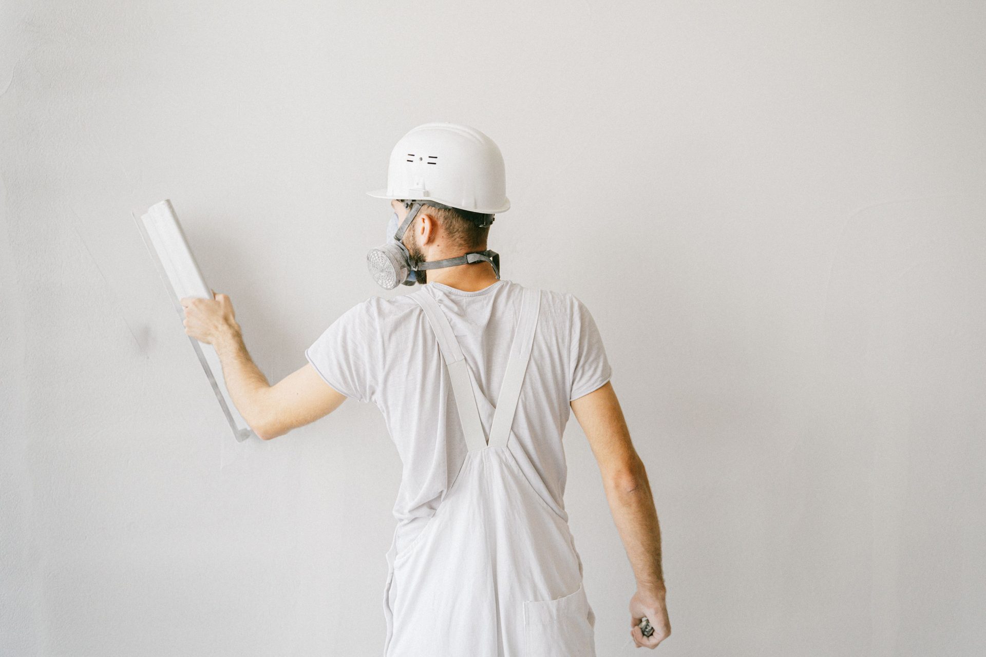 A plasterer seen from behind, in a hard hat and mask, smoothing a white wall.